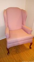 Salmon-colored wingback chair with wooden legs on hardwood floor against a wall.