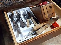 Drawer with stainless steel forks, spoons, and knives with light wooden handles plus a wooden knife block and some kitchen tools.