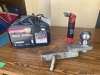 Motomaster 10 amp battery charger with cables and voltage meter visible, plus torch lighter and trailer hitch arranged on a wooden surface