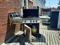 Propane BBQ grill outside, showing front with open bottom door and propane tank inside, grill lid open, control knobs visible.