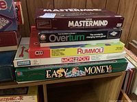 Stack of board games including Mastermind, Overturn, Rummoli, Monopoly, and Easy Money on a wooden surface.