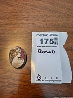 Photo of oval cameo brooch displaying the profile of a woman in cream relief against reddish background with decorative frame, placed on wooden surface with lot tag.
