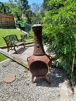 Front and side view of rusty red cast iron chimenea standing on gravel in yard with benches and greenery in background.