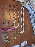 Photo shows an overhead view of various costume jewelry items laid out on a wooden surface. Items include multiple beaded necklaces in white and blue, bracelets with stones, ornamental silver-tone wide bracelet, various pins, brooches, and pendants. A thin gold-tone chain necklace is also visible.