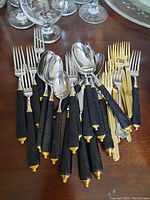 Cutlery set with black textured handles and gold accents, including forks, spoons, and knives arranged on a wooden table.