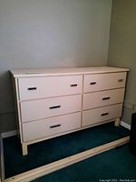 Photo of off-white wooden dresser with 6 drawers, showing front and part of side. Mixed drawer handles in different finishes and some scratches on surface.