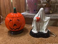 Photo showing both lamps: an orange Jack-o-Lantern and a white ghost lamp, both plugged in and standing on carpet.