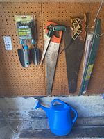 Photo showing four hand saws with various handles hanging on pegboard and blue plastic watering can on floor below.