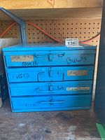 Blue steel hardware cabinet with four drawers, each labeled for contents such as bulbs and fuses, placed on a wooden surface against a pegboard wall.