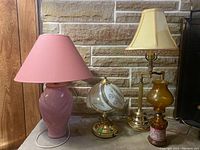 Four lamps on a wooden surface against a stone and wood wall background, including a pink ceramic lamp, brass lamp with white floral globe, brass adjustable lamp, and amber oil lamp.