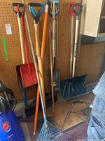 Photo showing several long-handled shovels including red plastic, green plastic, and metal varieties, along with blue metal rake and other tools leaning against a wall.