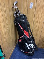 Full view of Founders Club golf bag and set of right handed golf clubs against wood panel wall.