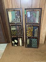 Two metal wall art panels leaning against a wood-paneled wall showing detailed abstract botanical and geometric patterns in greens and browns.