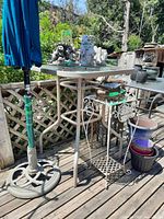 Photo of patio bar height table with beige metal frame, glass top, and umbrella stand base outdoors on wooden deck.