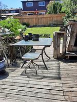 Outdoor wooden deck with metal framed glass top patio table of size 38x38x28 inches. Visible curved metal legs and a frosted glass top with some patio accessories on it.