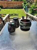 Photo of two black metal lanterns and one glossy black metal planter on glass patio table, outdoor background with grass and fence.