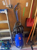 Full view of the blue Simoniz S1900 pressure washer with hose wrapped around the holder, placed against garage wall with tools nearby.