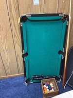 Photo showing small pool table with green felt surface and six pockets, standing upright against wood-paneled wall, with billiard balls in a wooden box and two black cues beside it.