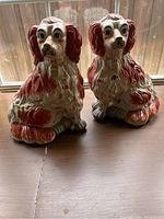 Pair of detailed ceramic spaniel dog figurines in rust-red and cream colors, both sitting poses on a wood surface in front of a window.