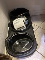 Photo showing three black Farberware slow cookers lined up, each with removable inner pots and clear lids. The slow cookers are brand new with protective packaging inside one pot.