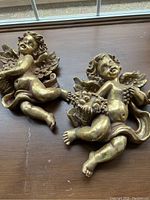 Photo showing two gold cherub angel sculptures placed on a wooden surface with detailed view of both figures, wings, and grapes.