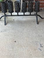 Six black iron candle holders mounted on horizontal stand placed on a concrete floor.