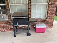 Outdoor black Expert grill on legs and wheels with side shelf, placed next to a red and white wheeled cooler against a brick wall.