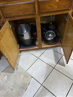 Open wooden cabinet showing several stacked metal pots and pans inside on tiled floor.