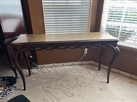 Photo of a tall wooden sofa table with curved legs and carved decorative trim along the front, placed near a window on a dirty floor.