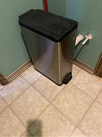 Photo of a rectangular stainless steel trash can with a black lid and foot pedal sitting on tiled floor by a green wall and baseboards.