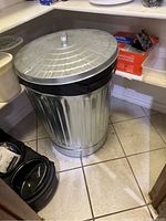 Metal kitchen trash can with removable lid placed on tiled floor in a corner, showing ribbed round body and lid handle.