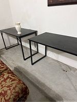 Two black folding tables with metal legs placed side by side on a concrete floor against a white wall. One table has a ceramic vase on it.