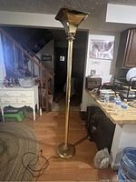 Full view of tall brass floor lamp in room setting showing column, brass finish, square top shade and round base with electrical cord on floor.