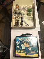 Box containing the G.I. Joe figure dressed in camouflage with military accessories alongside the Adam-12 lunch box featuring police officers artwork on lid.