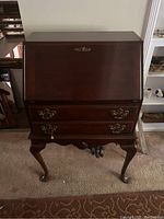 Closed view of the dark wood slant top desk featuring brass handles and cabriole legs.