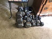 Photo showing row of hexagonal metal dumbbells in varying sizes placed on carpeted floor near furniture.