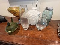 Photo showing seven glass collectibles including bowls, vases, covered jar, lidded dish, and sugar shaker on wooden surface