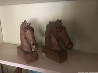 Two brown carved horse head bookends on a shelf, one with visible damage at base.