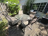 Outdoor patio set on wooden deck, includes round glass table and four chairs with brown metal frames and mesh fabric, arranged around table