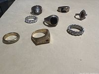 Seven assorted rings displayed on a beige surface with a variety of stones and metal finishes.