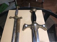 Top view of two decorative swords on a table, showing detailed hilt and scabbard of the narrow-bladed sword and crossguard of the wide-bladed sword