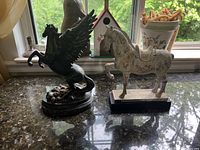 Two horse figurines on countertop by window: a dark metal Pegasus with wings and a distressed white horse on a black rectangular base.