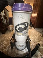 Front view of the white cylindrical Super Coach backpack vacuum cleaner with purple top, showing the main body with the logo and the attached hose on the floor.