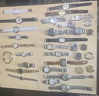 Full view of 28 men's and women's watches arranged on a tan surface with a range of band styles including metal, leather, rubber, and mesh.