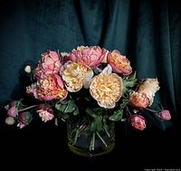 Front view of silk peony floral arrangement showing full bloom flowers in pink and cream with green leaves in a clear glass vase against a dark backdrop.