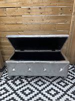 Front-facing photo of silver upholstered storage bench with top open, showing black storage compartment and cabriole legs.