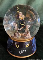 Front view of water globe showing three figurines inside a clear sphere, base with cat eyes and the word 'CATS'.
