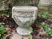 View of concrete planter sitting in garden soil surrounded by plants, showing front side with Greek key rim and fruit detail.