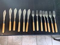 Full set of six fish knives and six forks on dark table, showing bone handles and silver or silver-plated engraved blades on knives, one fork different from others