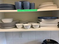Shelf displaying stacks of dishware including white plates, white bowls, blue and white ceramic plates, gray cups, and rectangular melamine plates.